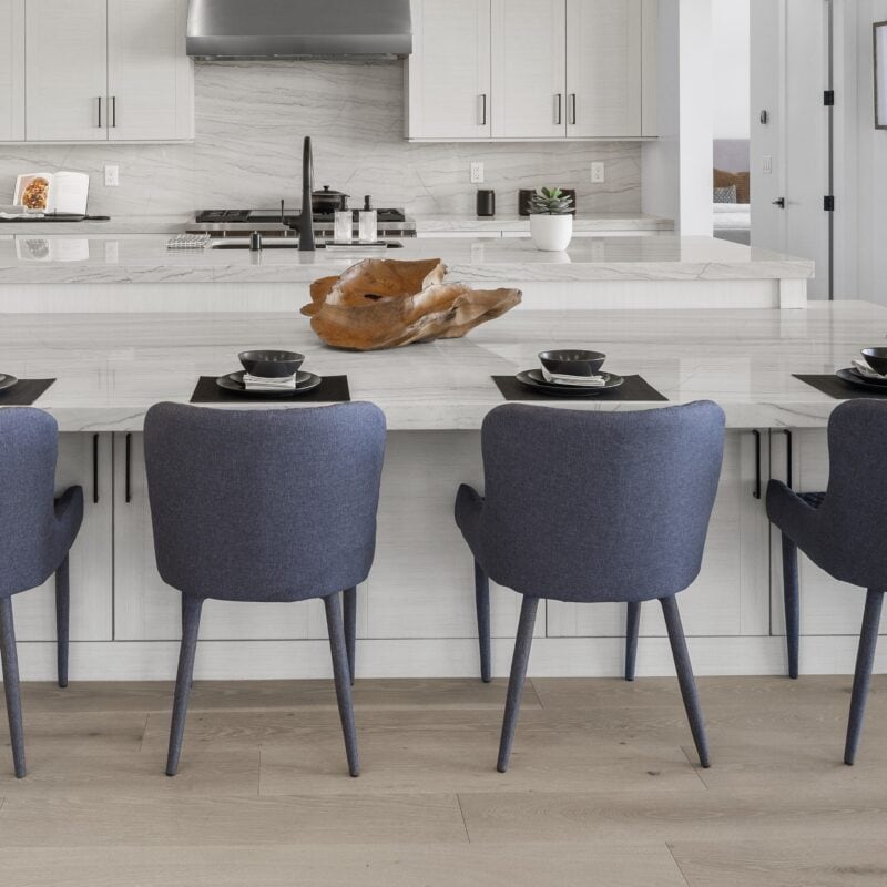 Modern dining room with a large white table, eight blue upholstered chairs, and minimalist kitchen i.